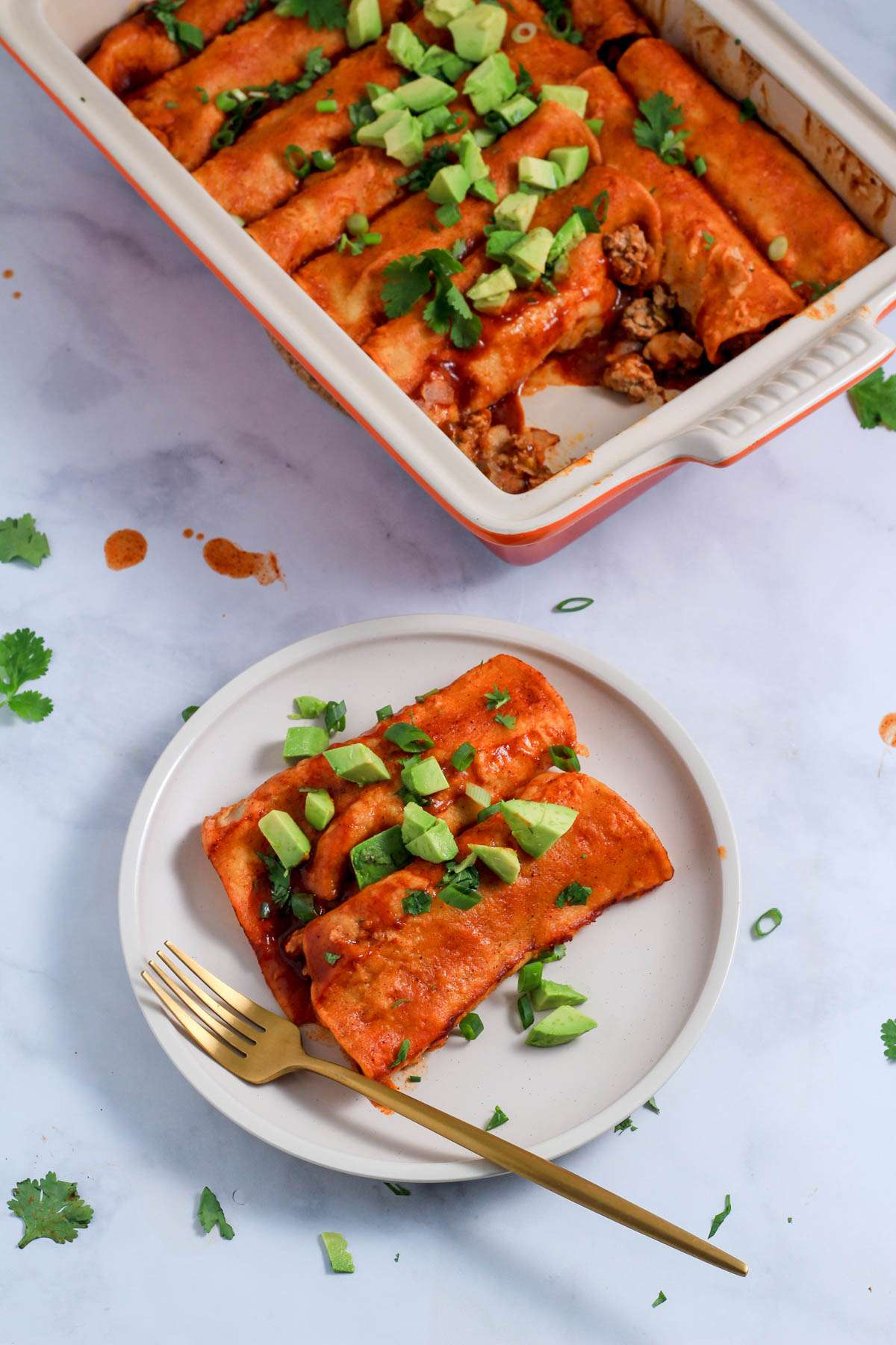 A top down picture of ground turkey enchiladas on a white plate topped with cilantro, avocado, and green onion with a gold fork on the plate in front of the baking pan of enchiladas.