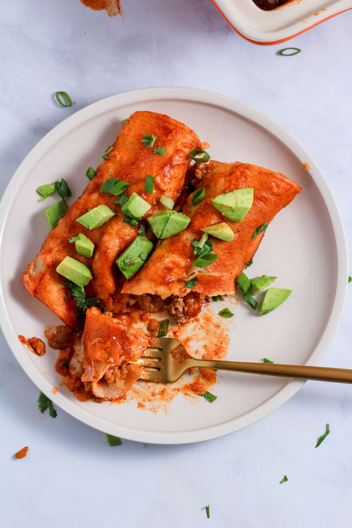 A white plate with two ground turkey enchiladas topped with avocado and cilantro with a gold fork on a white counter.