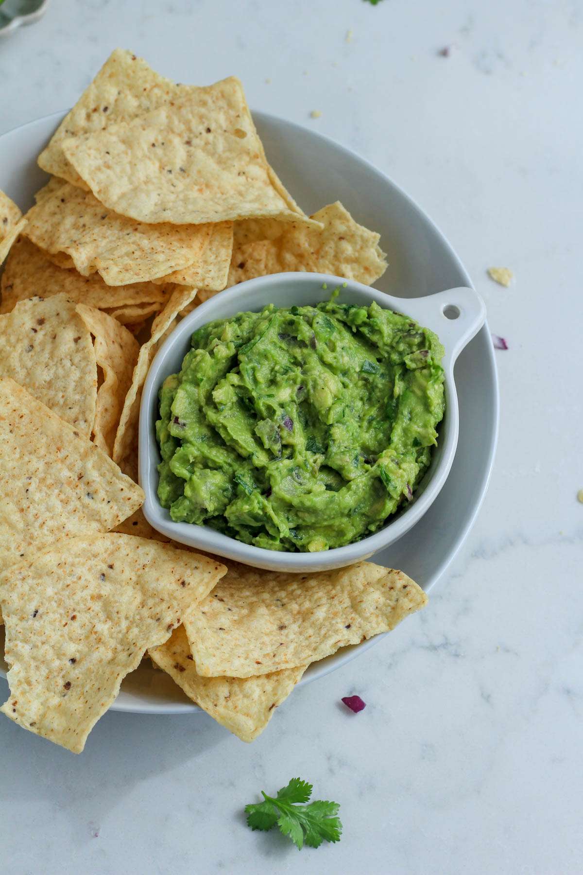 A small white bowl filled with vegan guacamole inside a larger white bowl filled with tortilla chips on a white counter.