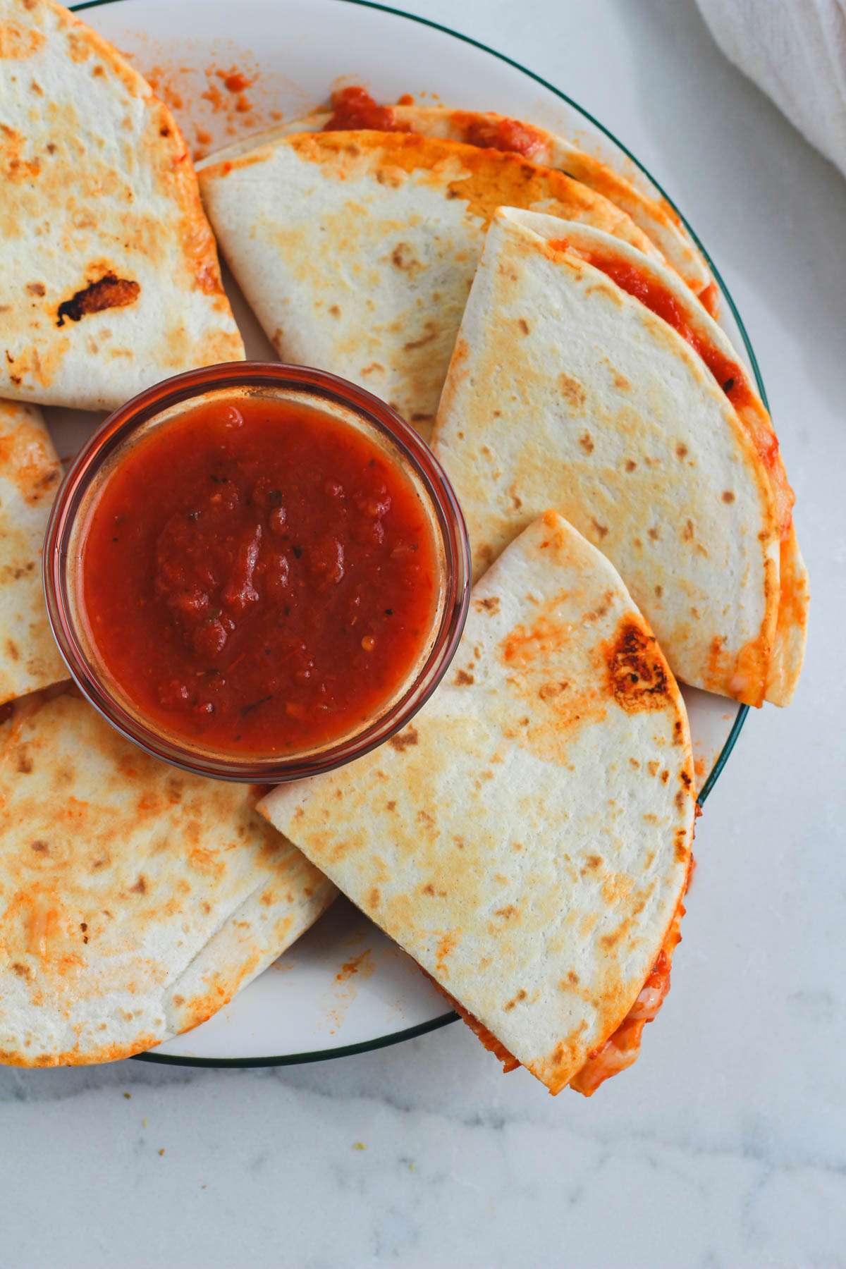 A top down image of dairy-free pizzadillas on a white and green plate with a bowl of marinara sauce in the middle for dipping.