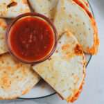A white plate with a green rim and wedges of dairy-free pizza quesadillas around a small bowl of marinara.