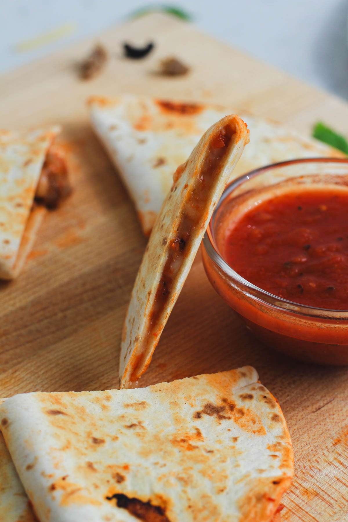 A wooden cutting board with wedges of pizza quesadilla next to a small bowl of marinara sauce.