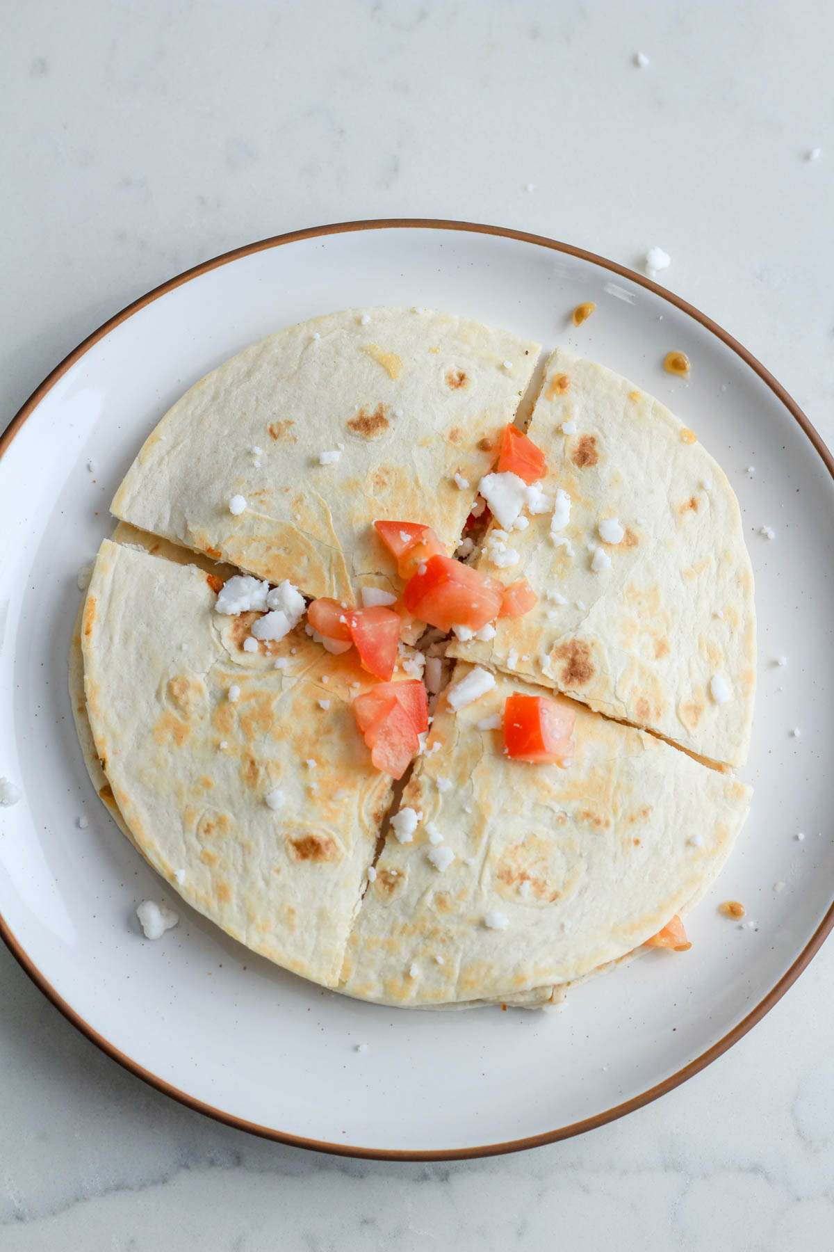 A white plate with a brown rim topped with a Greek chicken quesadilla garnished with Greek feta crumbles and diced tomatoes.