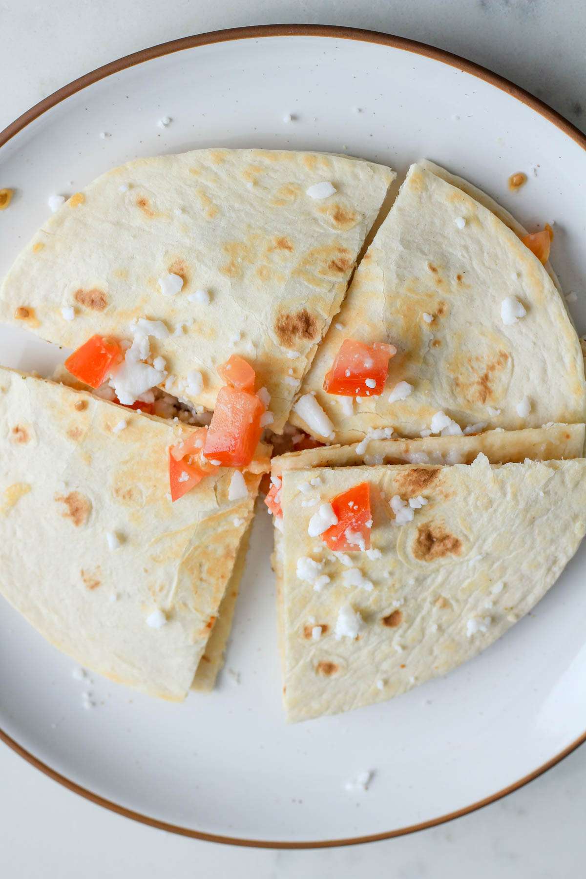 Wedges of a Greek chicken quesadilla topped with vegan feta and tomatoes on a white and brown plate.