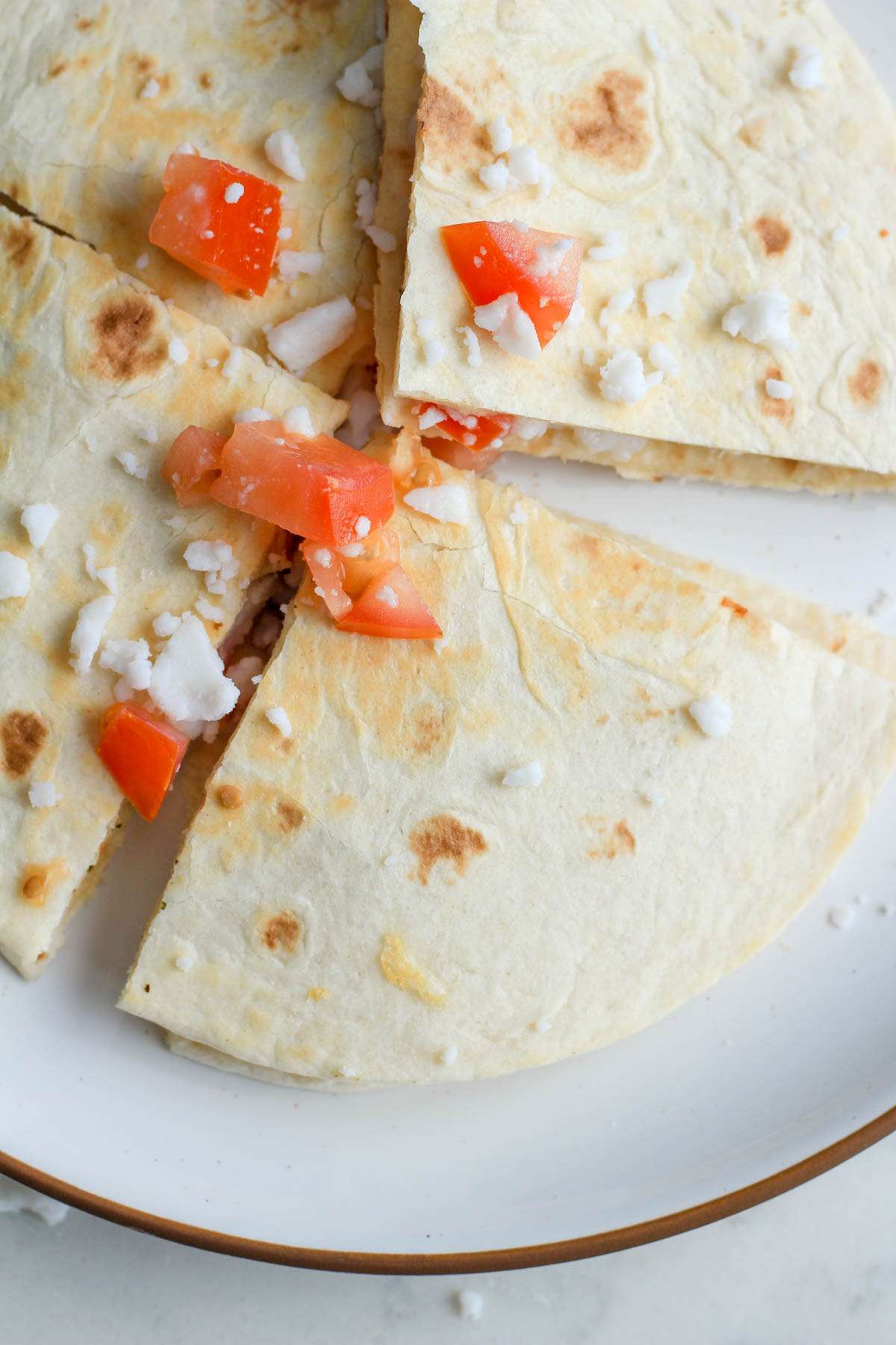 A white plate with a brown rim and a wedge of a Greek chicken quesadilla topped with Roma tomatoes and vegan feta.