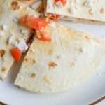 A wedge of Greek chicken quesadillas topped with vegan feta and tomatoes on a white plate with a brown rim.