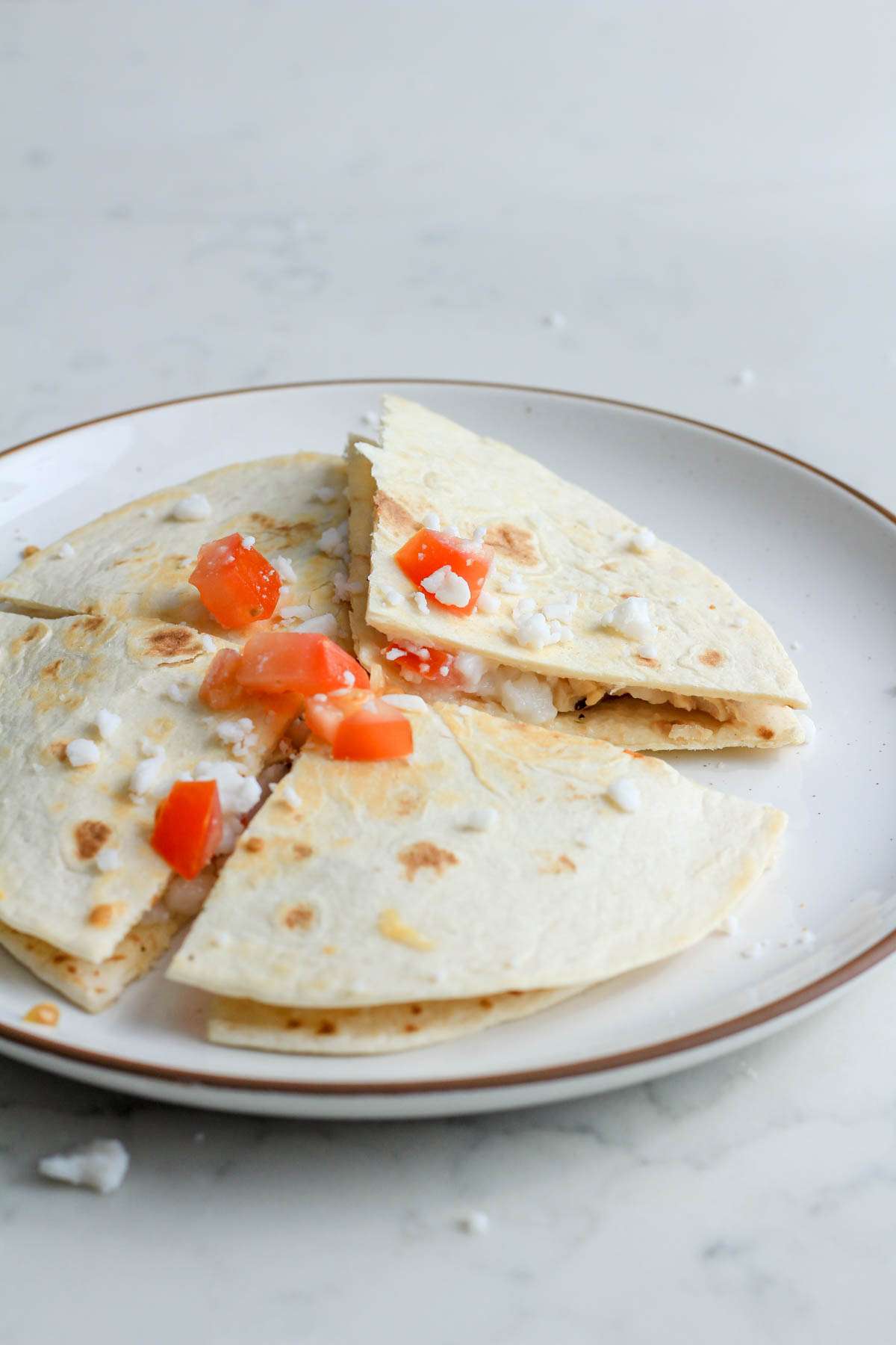 From the side, a white plate with a brown rim with a dairy-free greek chicken quesadilla topped with roma tomatoes and vegan feta crumbles.