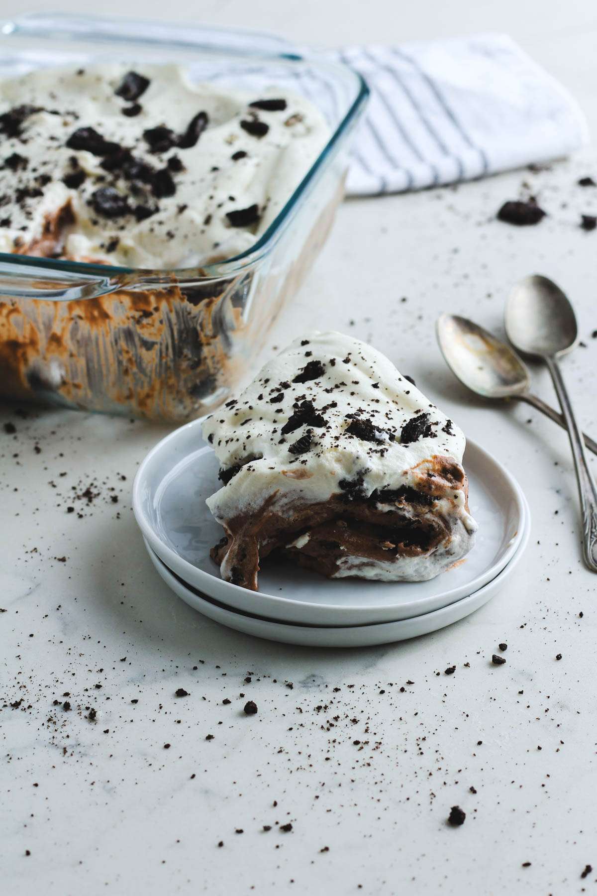 A white plate stack with a scoop of Oreo ice box cake with two silver spoons in the back right.