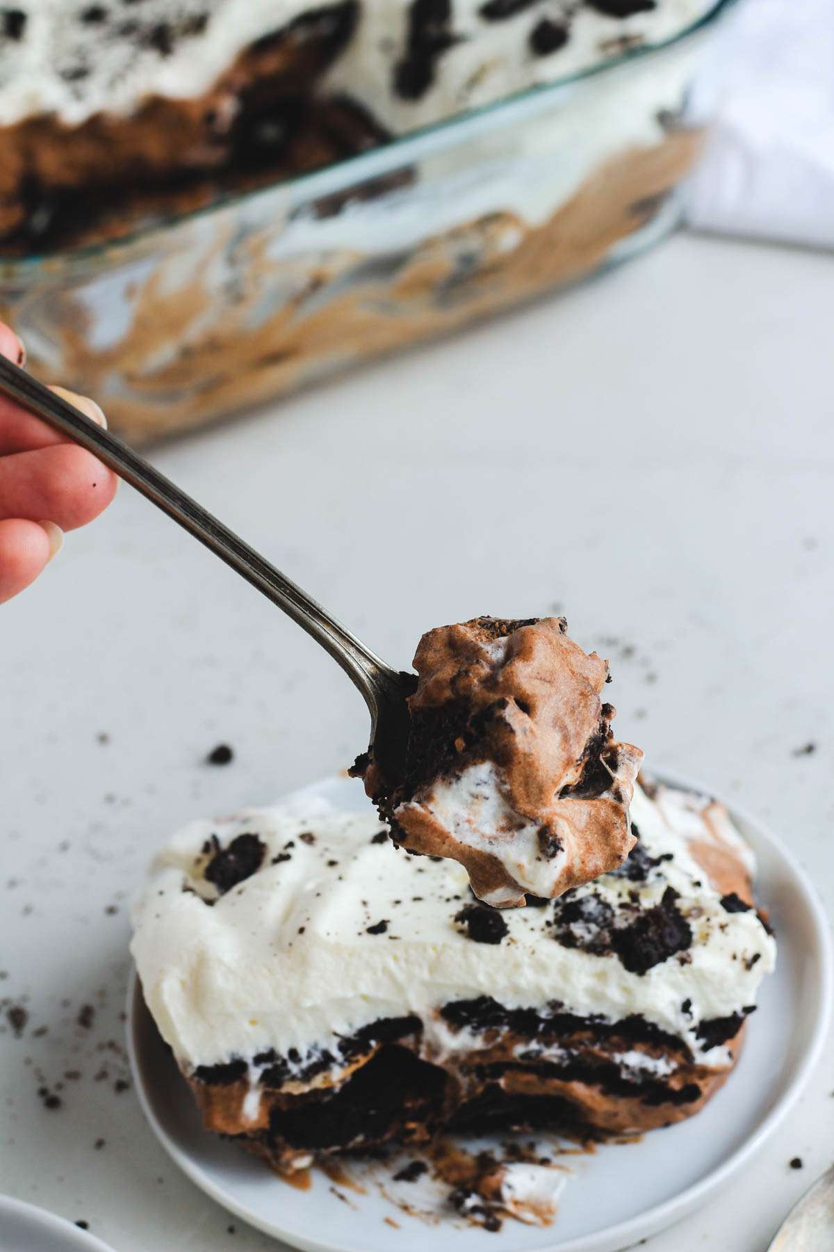 A hand holding a spoon with a scoop of Oreo ice box cake over a small white plate with a serving of dairy-free Oreo cake.
