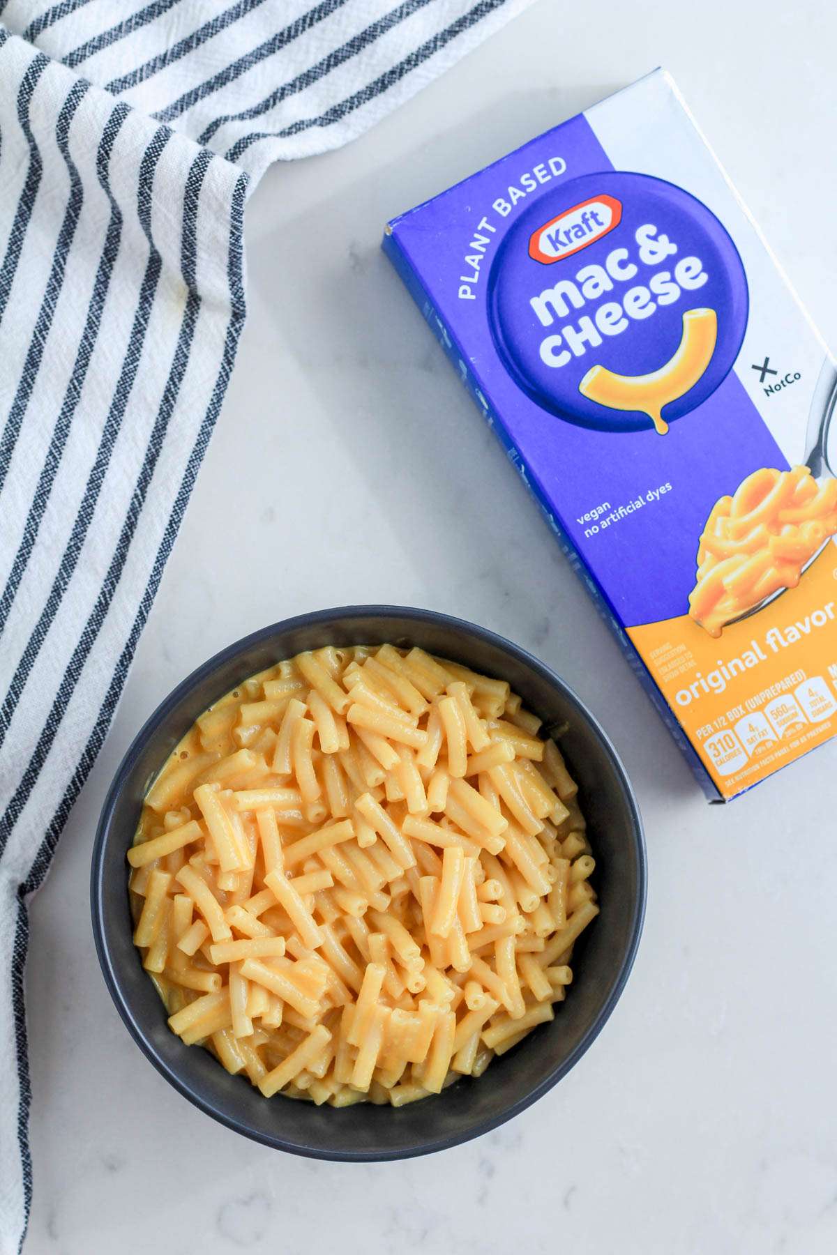 A bowl of original flavor vegan mac and cheese next to the box on a white counter with a blue and white striped dish towel.