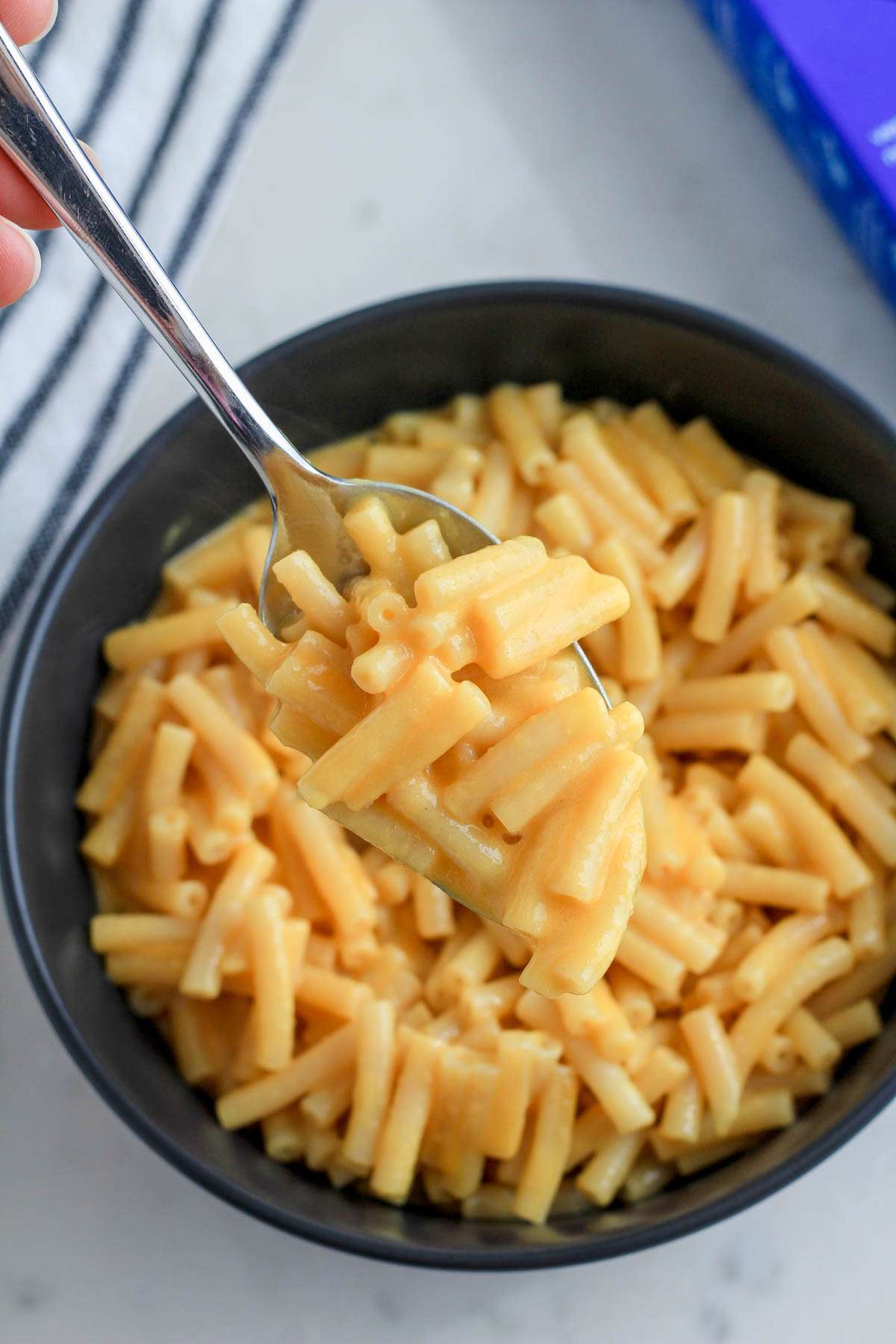 A silver spoon with a scoop of the original flavor plant based Kraft mac and cheese over a bowl of the mac and cheese.