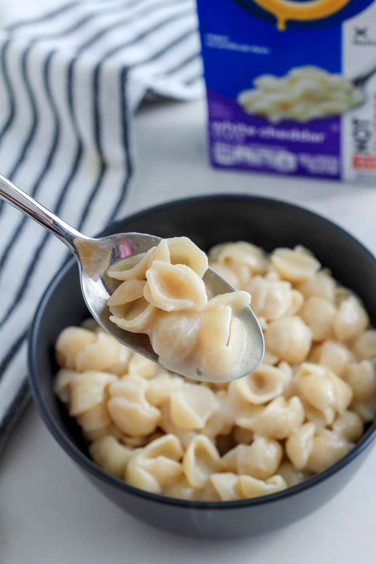 A spoon with a scoop of the white cheddar style plant based Kraft mac and cheese hovering over the bowl on front of the Kraft Not Cheese box.