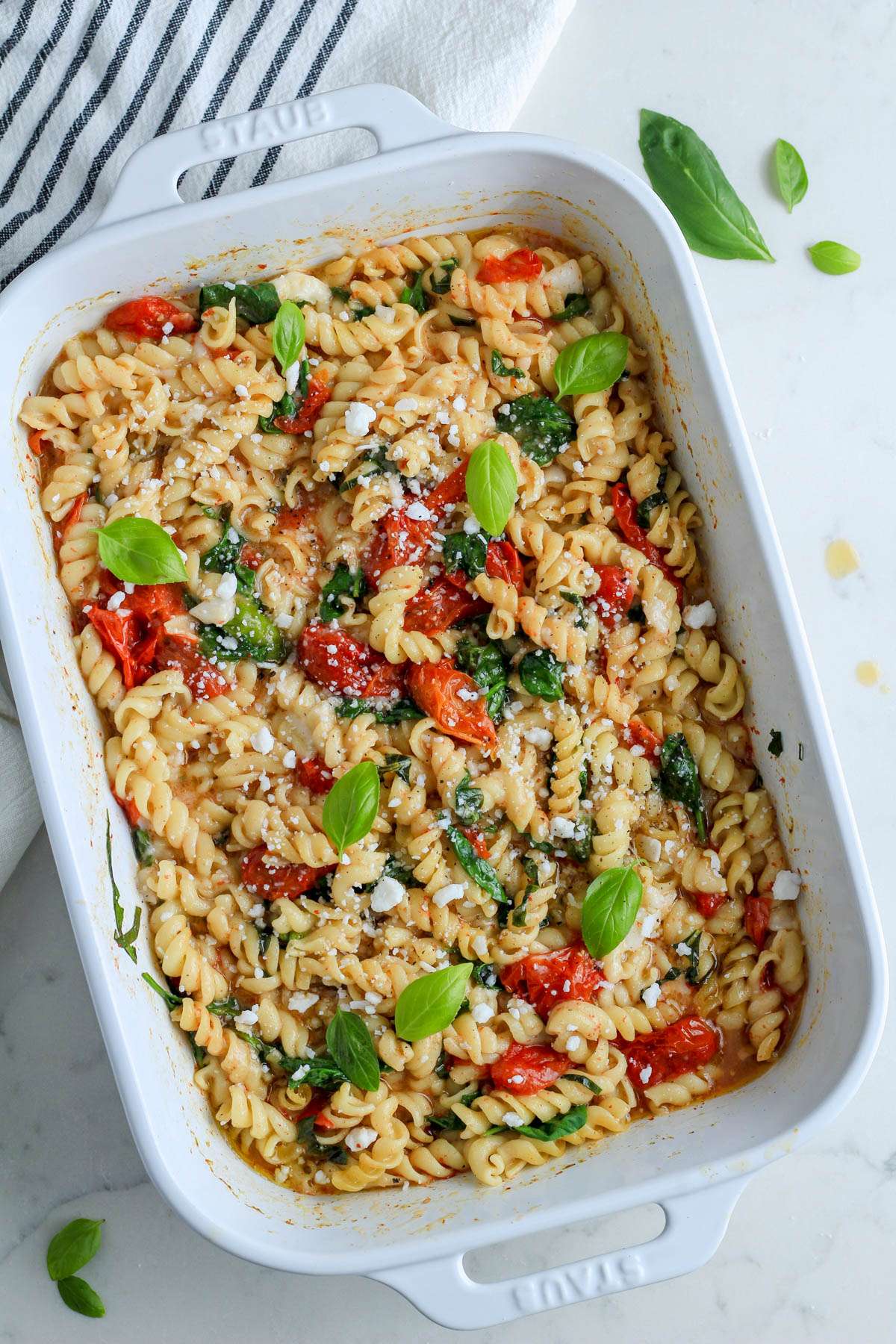 A top down photo of vegan baked feta pasta in a white baking dish on a white counter with a blue and white striped dish towel.