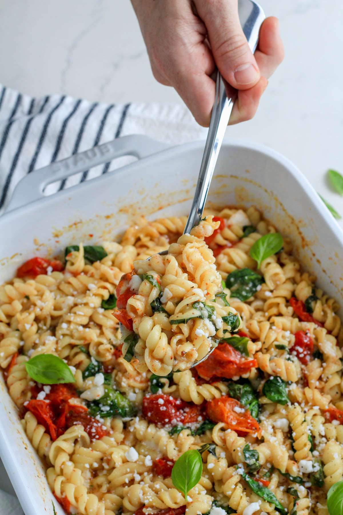A hand holding a silver spoon with a heaping scoop of vegan baked feta pasta over a white baking dish with more baked pasta.