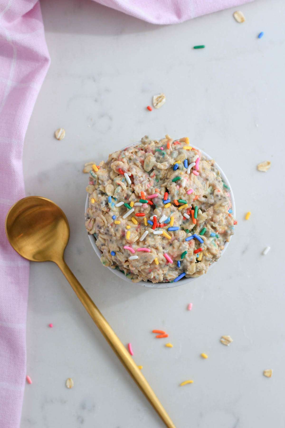 A top down photo of sprinkle covered vegan overnight oats with vanilla protein powder on a white counter with a gold spoon to the left.
