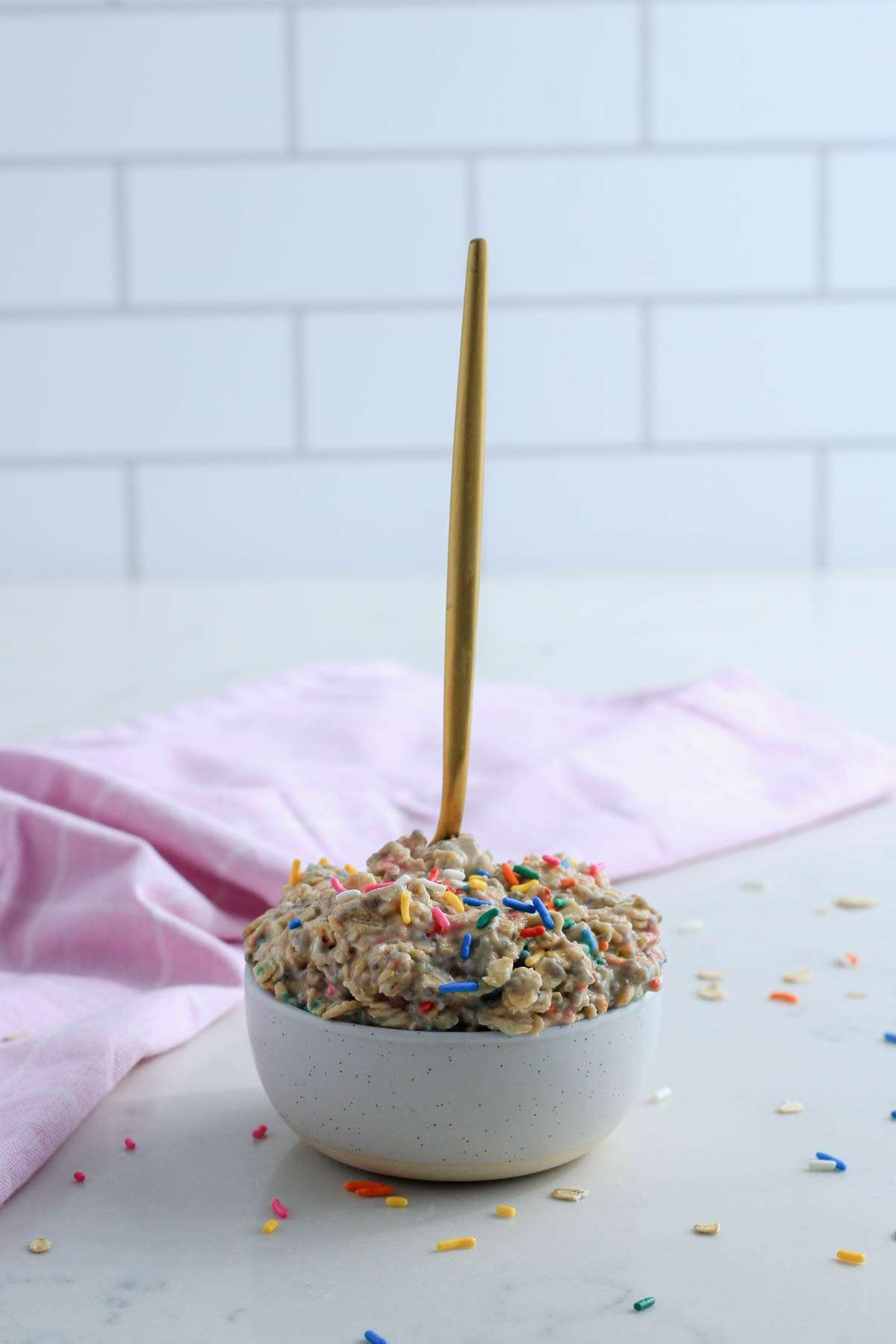 A gold spoon sticking out of the top of a white bowl filled with vegan overnight oats topped with sprinkles.