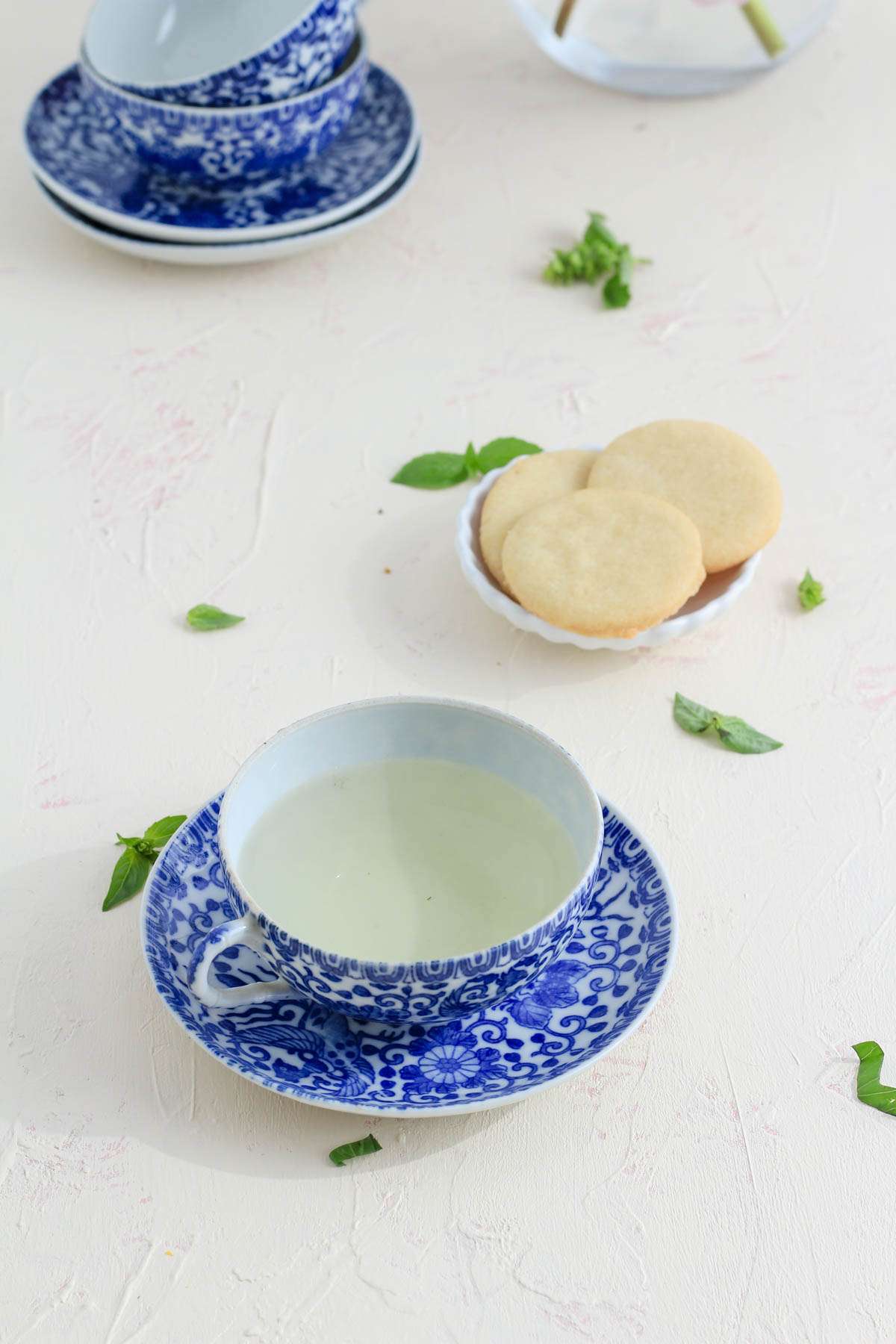 A white and blue tea cup with saucer on a white counter with a small dish of sugar cookies in the back right.