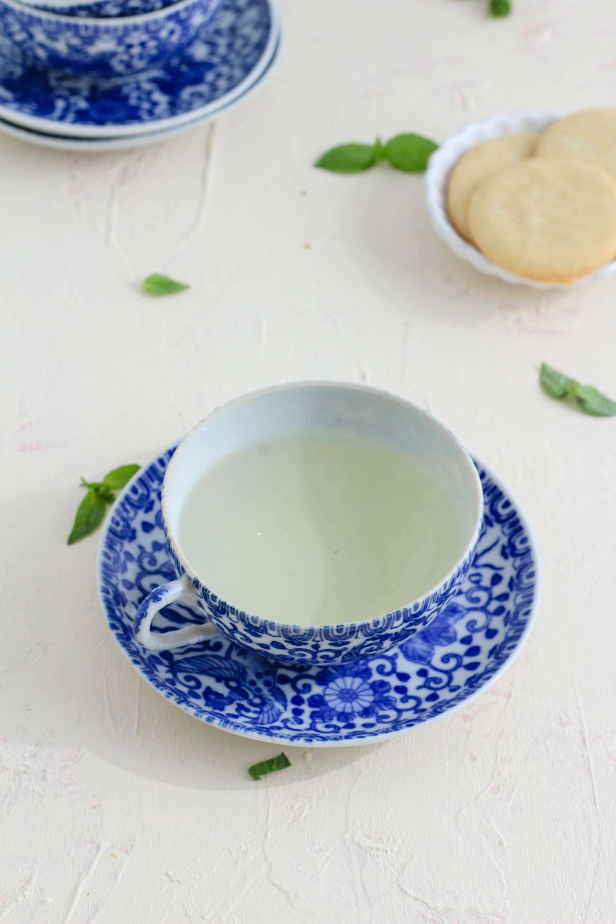 A cup of fresh basil tea on a blue and white saucer with fresh leaves scattered on the counter.