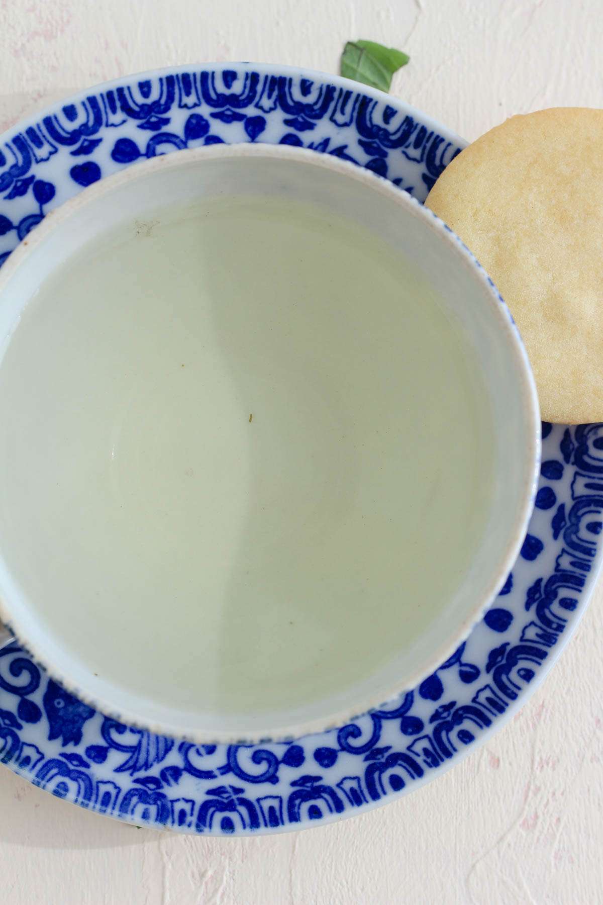 A top down image of fresh basil tea in a white and blue tea cup with a sugar cookie in the top right corner.