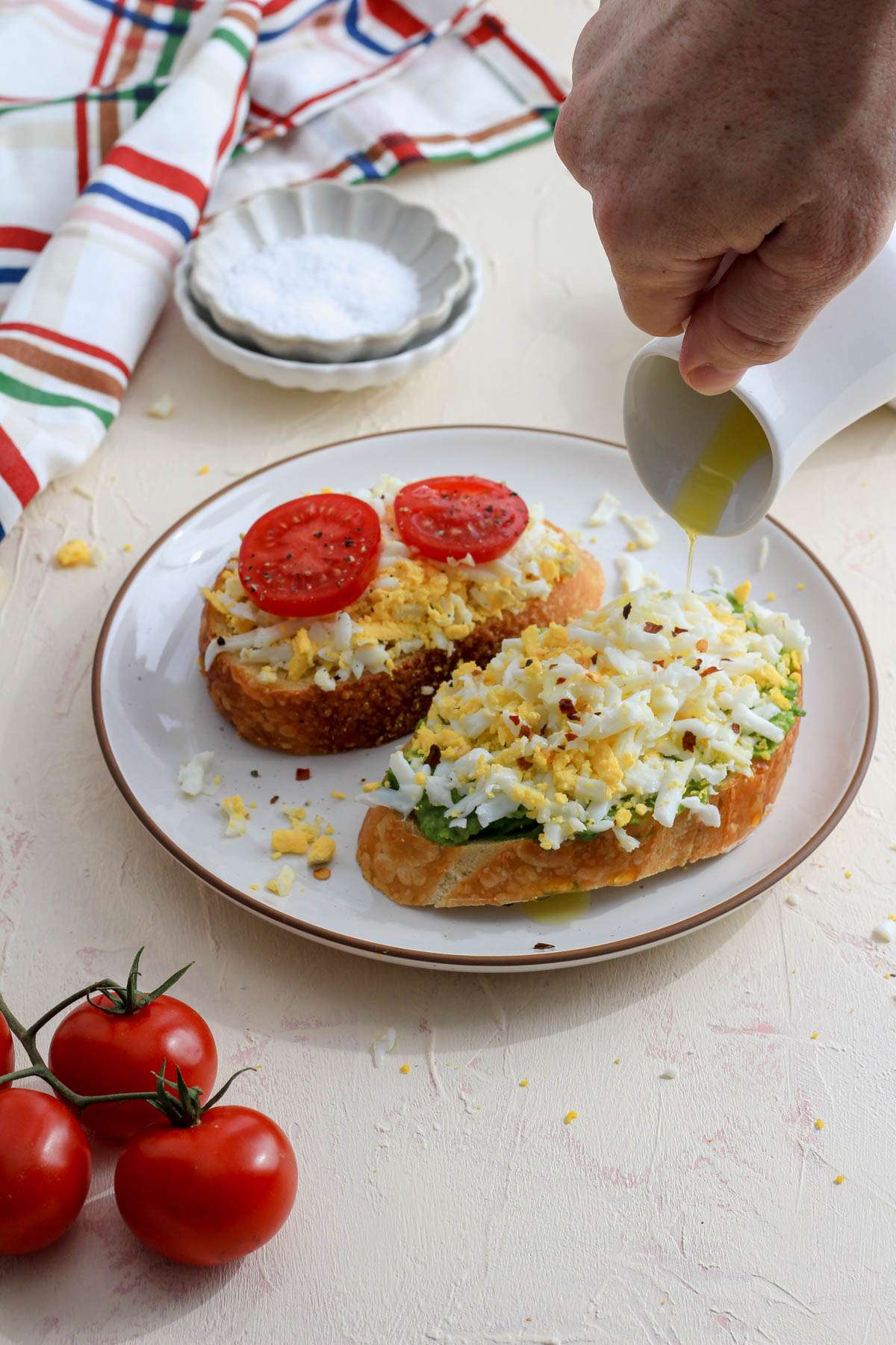 A white plate with a brown rim and two slices of grated egg toast with a hand drizzling olive oil on top.
