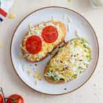 A white plate with a slice of grated egg on toast with two slices of tomato on the left and a slice of avocado toast with grated egg on the right.