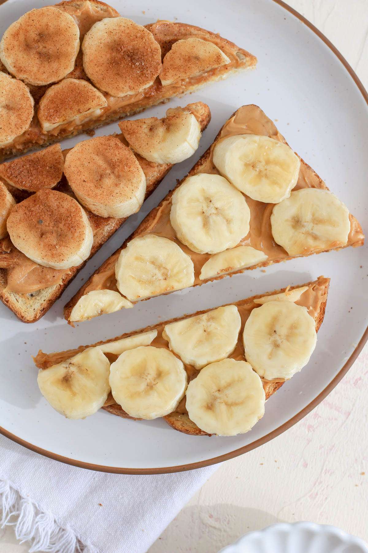 Four slices of peanut butter banana toast on a white plate with a brown rim.