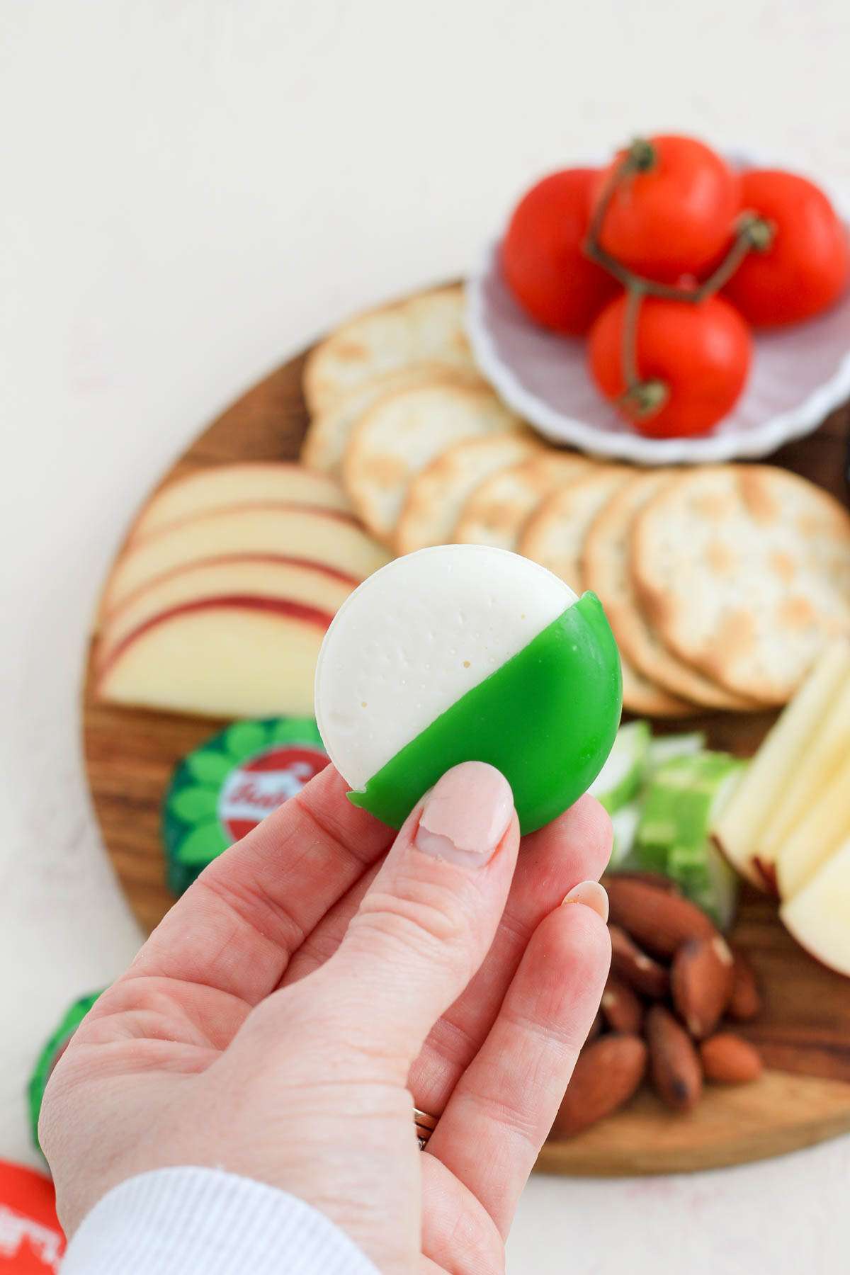 A hand holding a green wax wrapped piece of plant-based babybel cheese in front of a vegan cheese board.