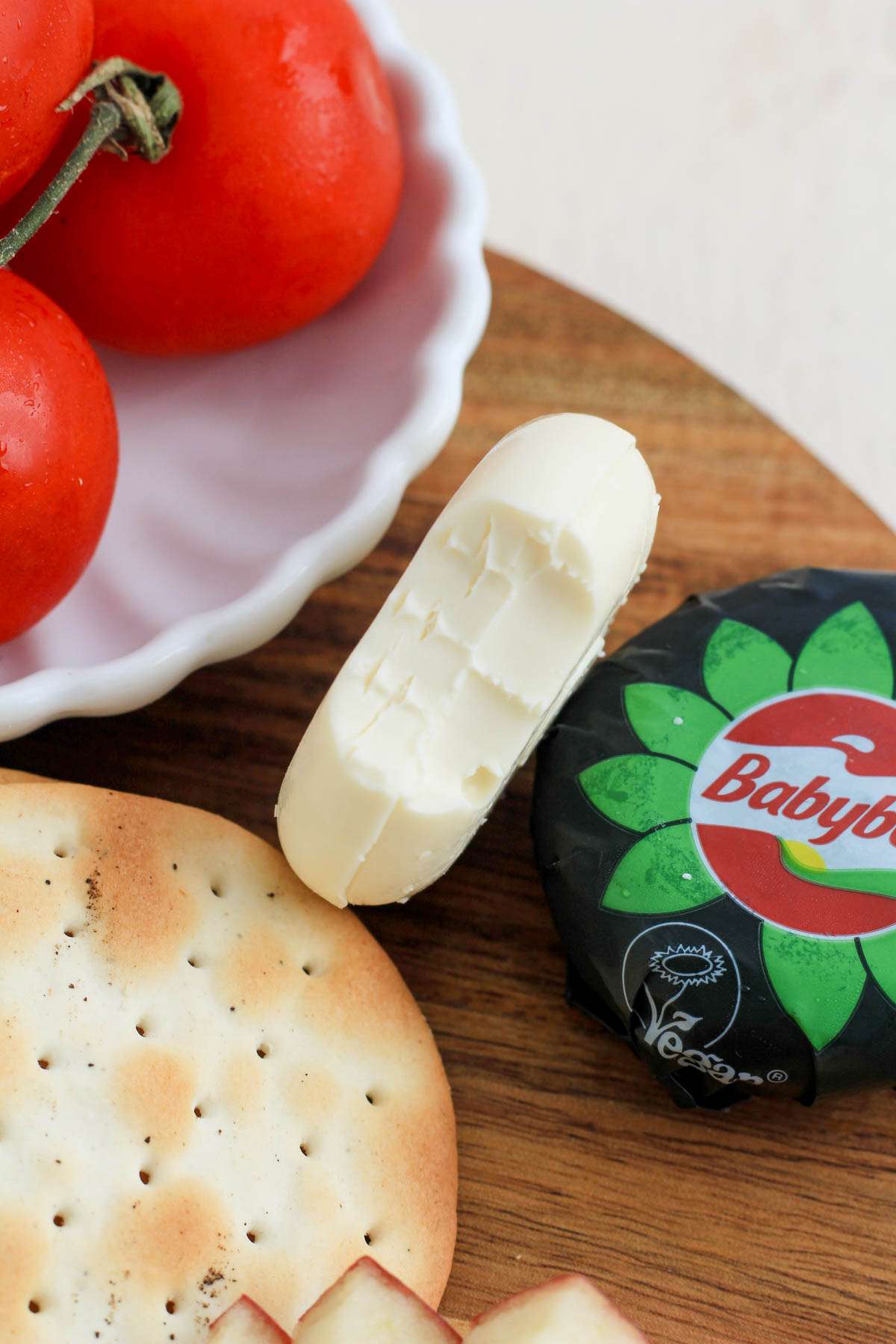 A bite taken out of a babybel cheese wheel on a wooden cutting board next to a cracker and tomatoes.