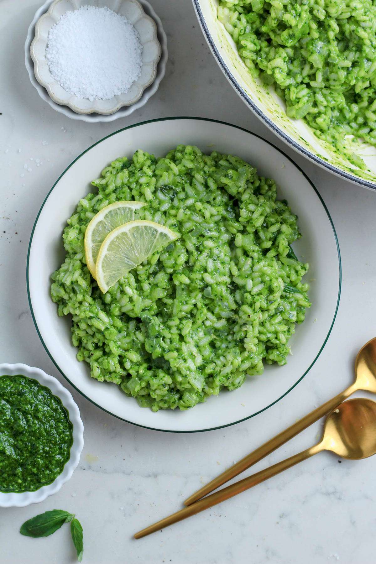A top down image of vegan pesto risotto on a green rimmed plate next to two gold spoons and a pinch bowl of salt.