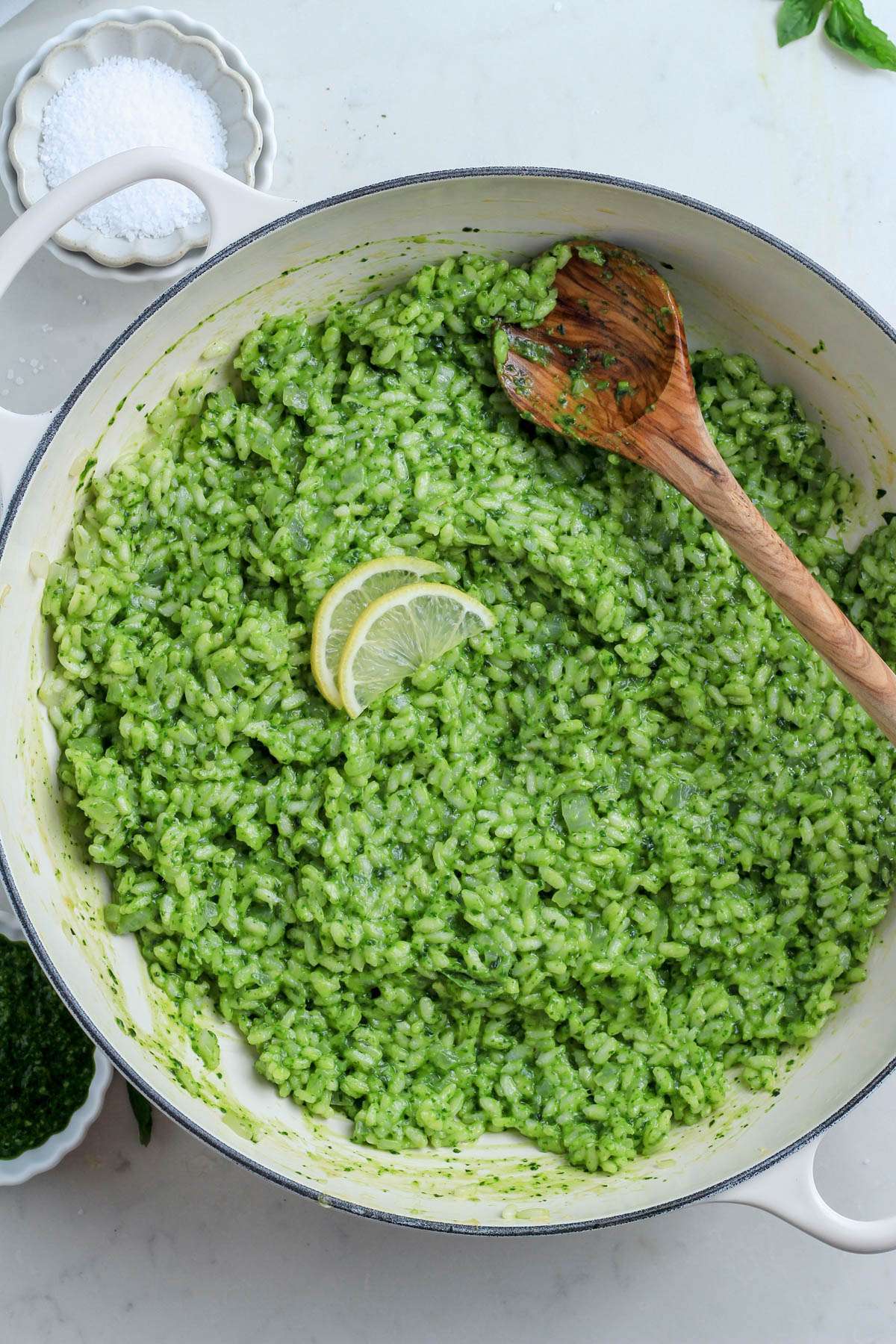 A deep dish skillet filled with spinach pesto risotto topped with slices of lemon and a wooden spoon in the top right of the skillet.
