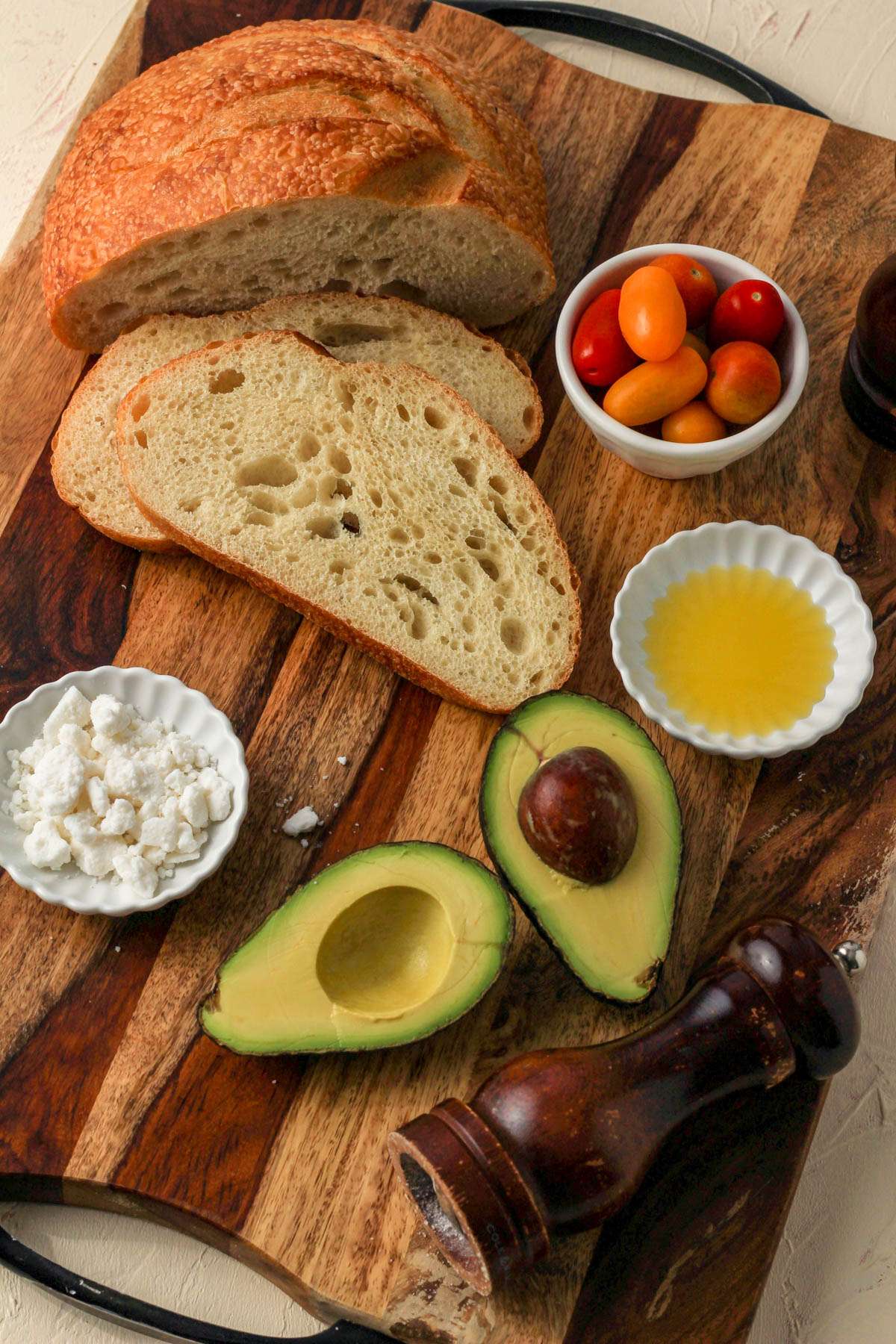 Ingredients for avocado toast on a wooden cutting board with bread, avocado, feta, and grape tomatoes.
