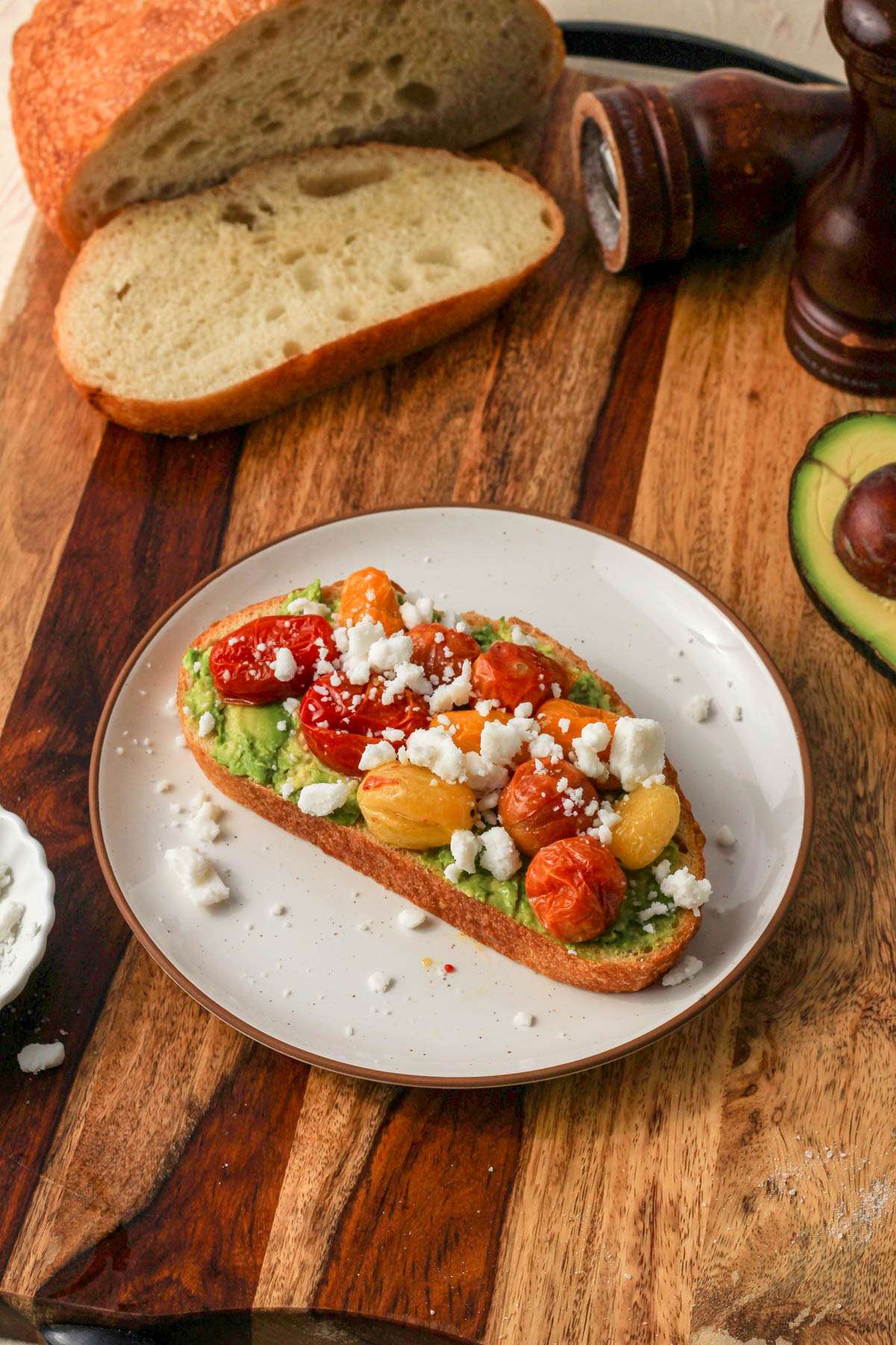 A small white plate with slices of toasted bread topped with smashed avocado and roasted grape tomatoes and vegan feta on a wooden cutting board.
