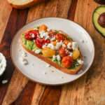 A small white plate with a slice of avocado toast with roasted tomatoes and feta.