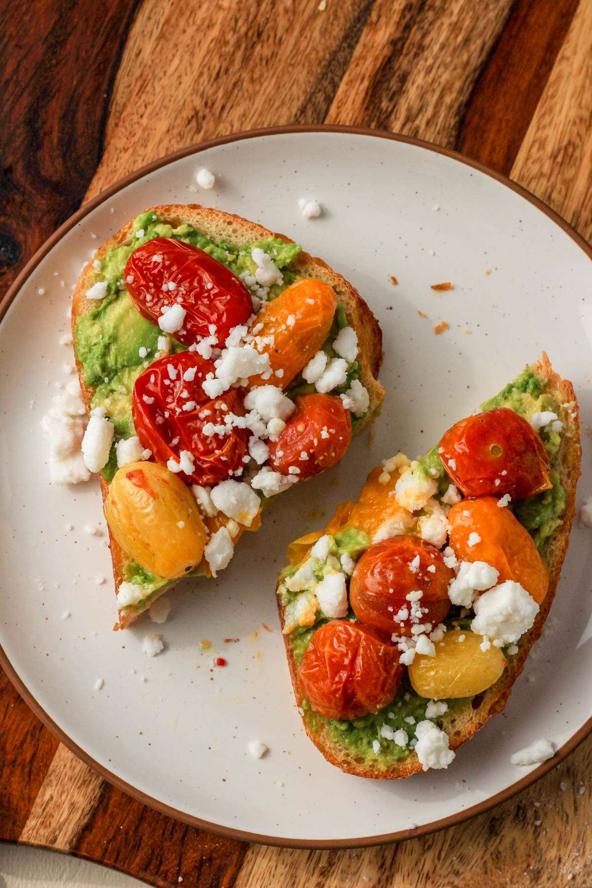 A top down picture of a white plate topped with avocado toast topped with roasted grape tomatoes and vegan feta.