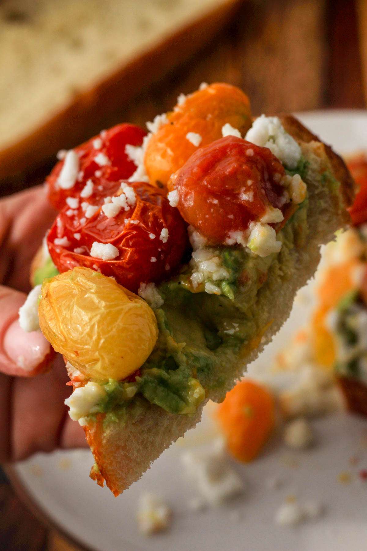A hand holding a slice of vegan avocado toast with roasted grape tomatoes and feta.