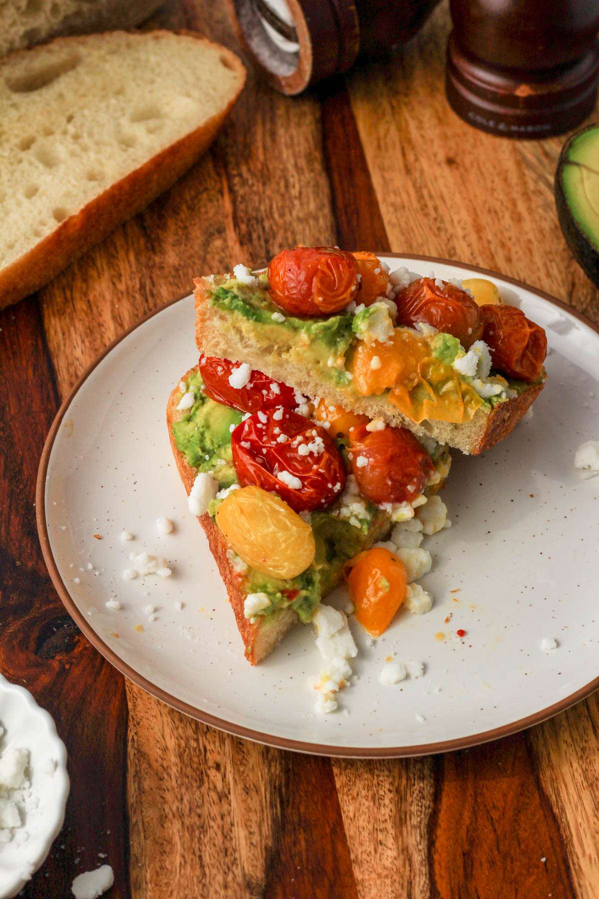 A slice of dairy-free avocado toast with roasted tomatoes and feta cut in half on a white plate.