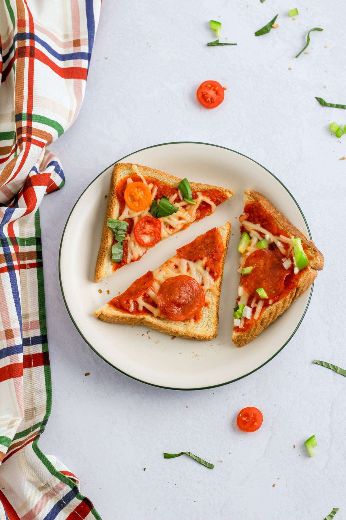 A green rimmed white plate with three different slices of dairy-free pizza toast on a white counter with a dish towel to the left.