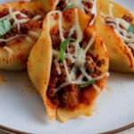 A close up of a stuffed shell with meat sauce topped with dairy-free cheese and fresh basil on a white plate.
