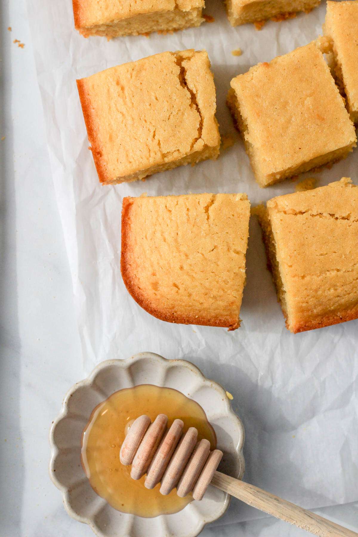 A white counter with a small bowl of honey in the bottom left with a parchment paper lined counter with slices of gluten-free cornbread on the parchment paper.