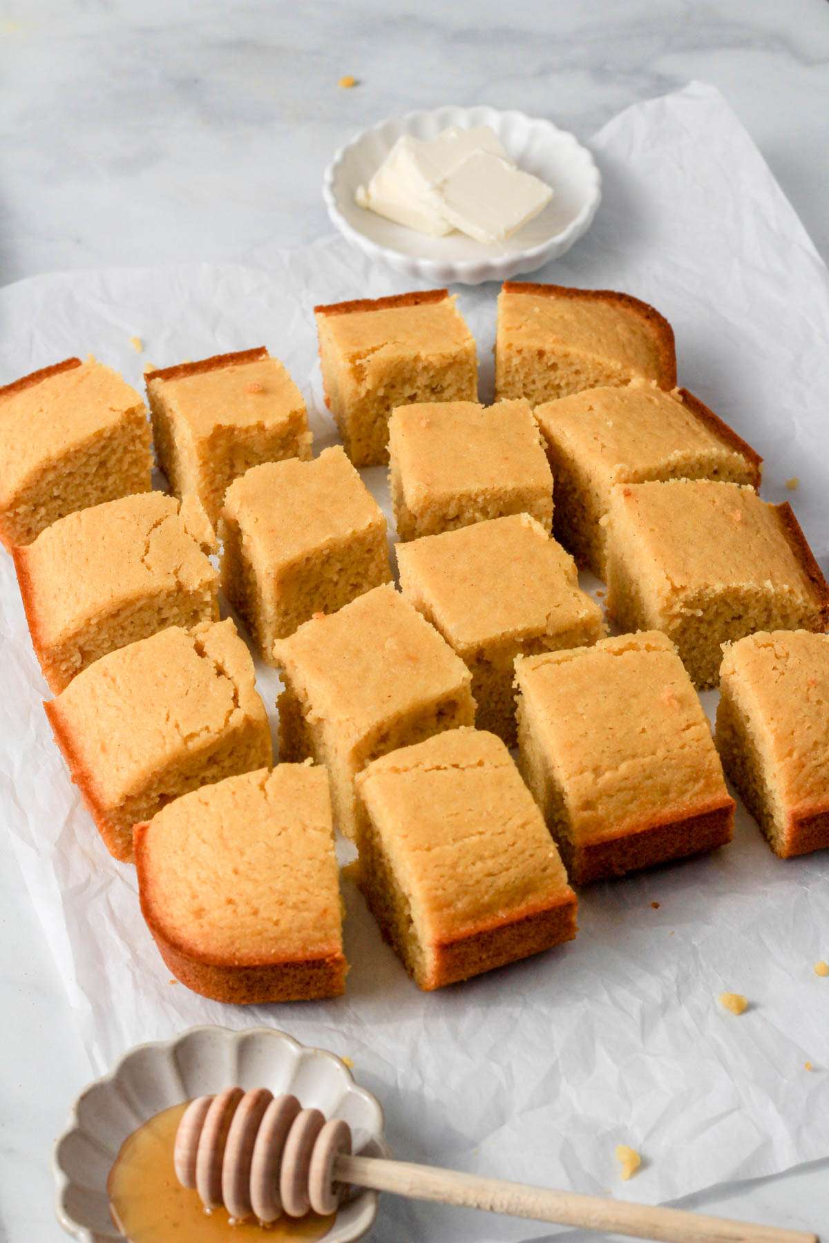 Gluten-free dairy-free cornbread on a slice of parchment paper with a bowl of honey and a bowl of butter in the corners.