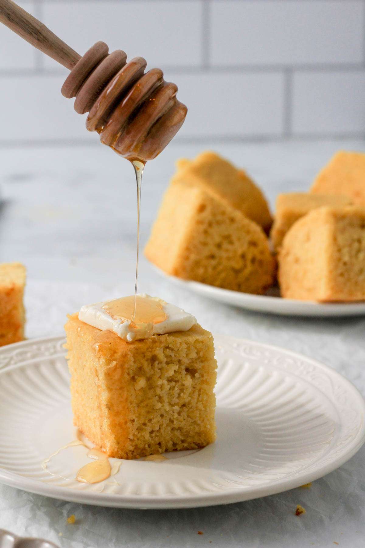 A small white plate with a slice of cornbread topped with vegan butter and a small spoon of honey dangling over the cornbread slice.