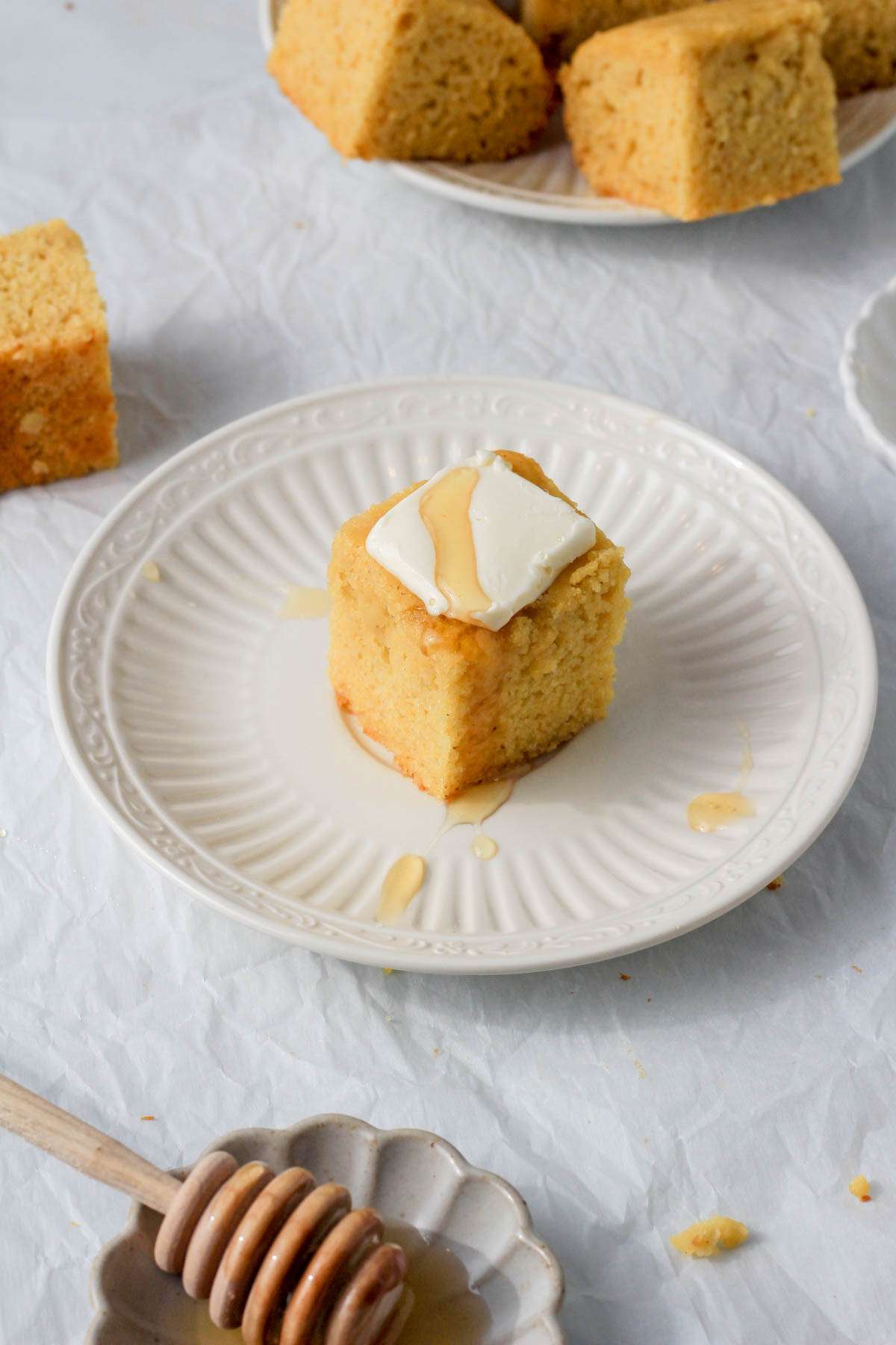 A small plate of dairy-free gluten-free cornbread with vegan butter and honey with a small plate of honey in the front left and a small plate of cornbread in the back right.