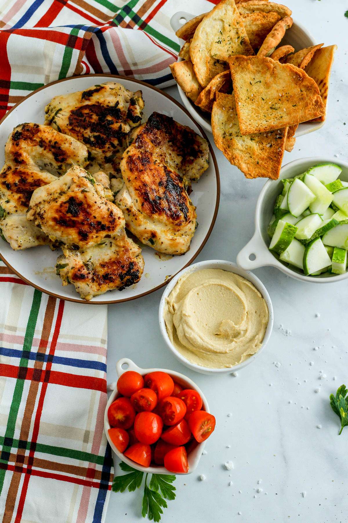 A white counter with a plate of chicken thighs, a bowl of pita chips, cucumber, hummus, and grape tomatoes.