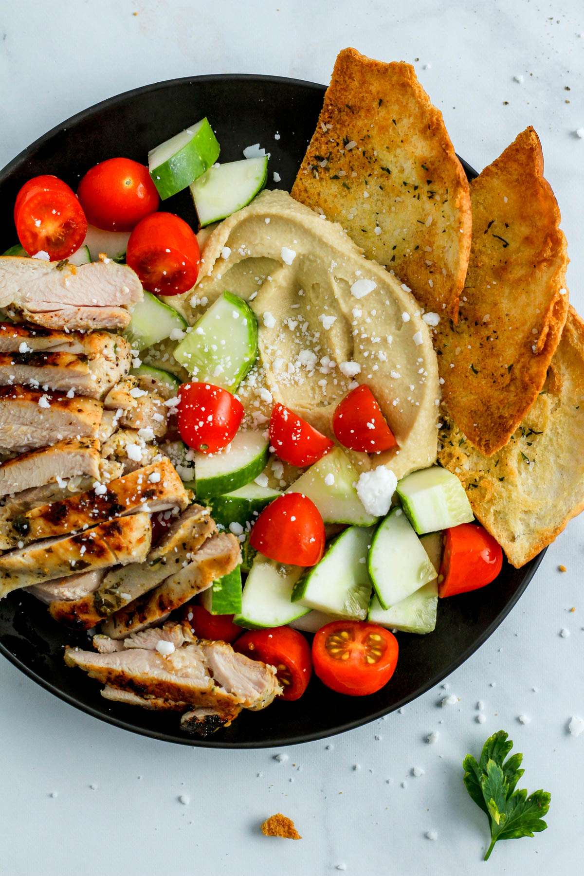 Greek chicken, cucumbers, tomatoes, hummus, and pita chips in a black plate on a white counter.