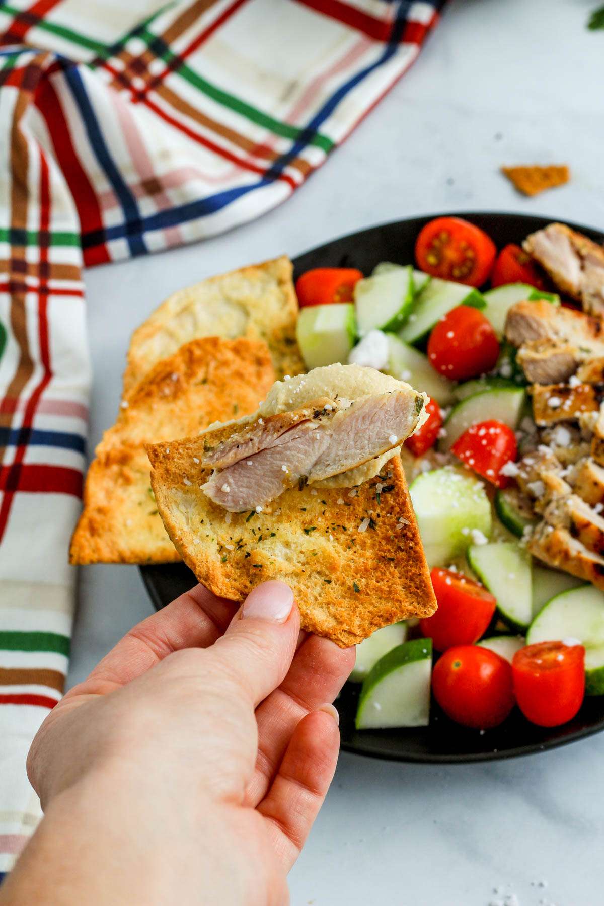 A hand holding a pita chip topped with hummus and chicken in front of a greek chicken hummus bowl.