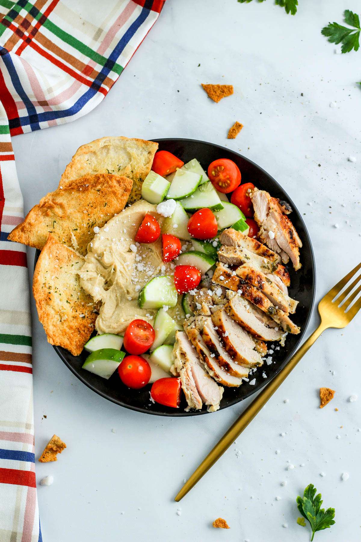 A black plate with grilled chicken, a cucumber and tomato salad, creamy hummus, and pita chips on a white counter next to a gold fork.
