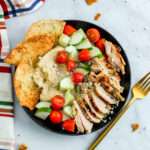 A greek chicken hummus bowl in a black bowl filled with chicken, cucumber and tomato, hummus, pita chips, and vegan feta on a white counter with a gold fork.