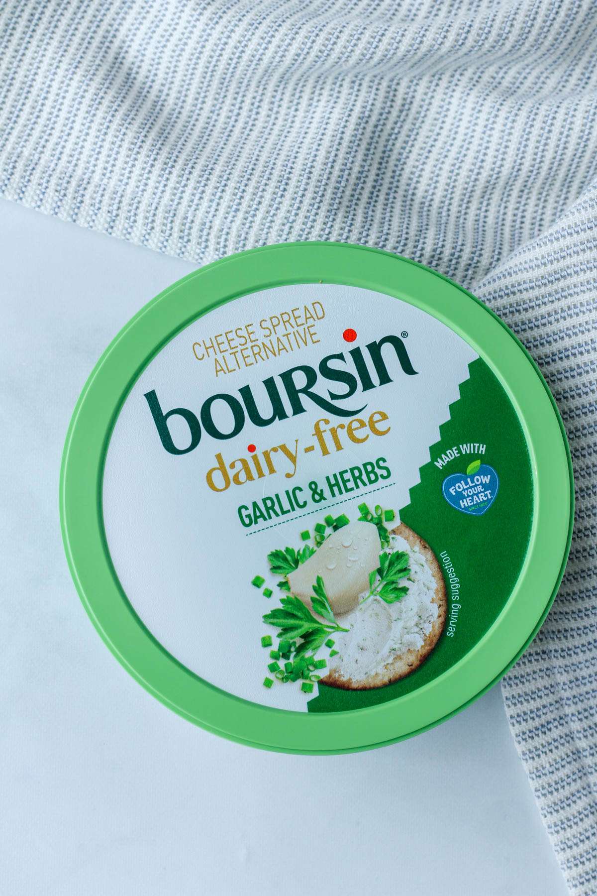 A green tub of Dairy-Free Boursin Garlic and Herb Cheese on a white counter with a dish towel to the top right.