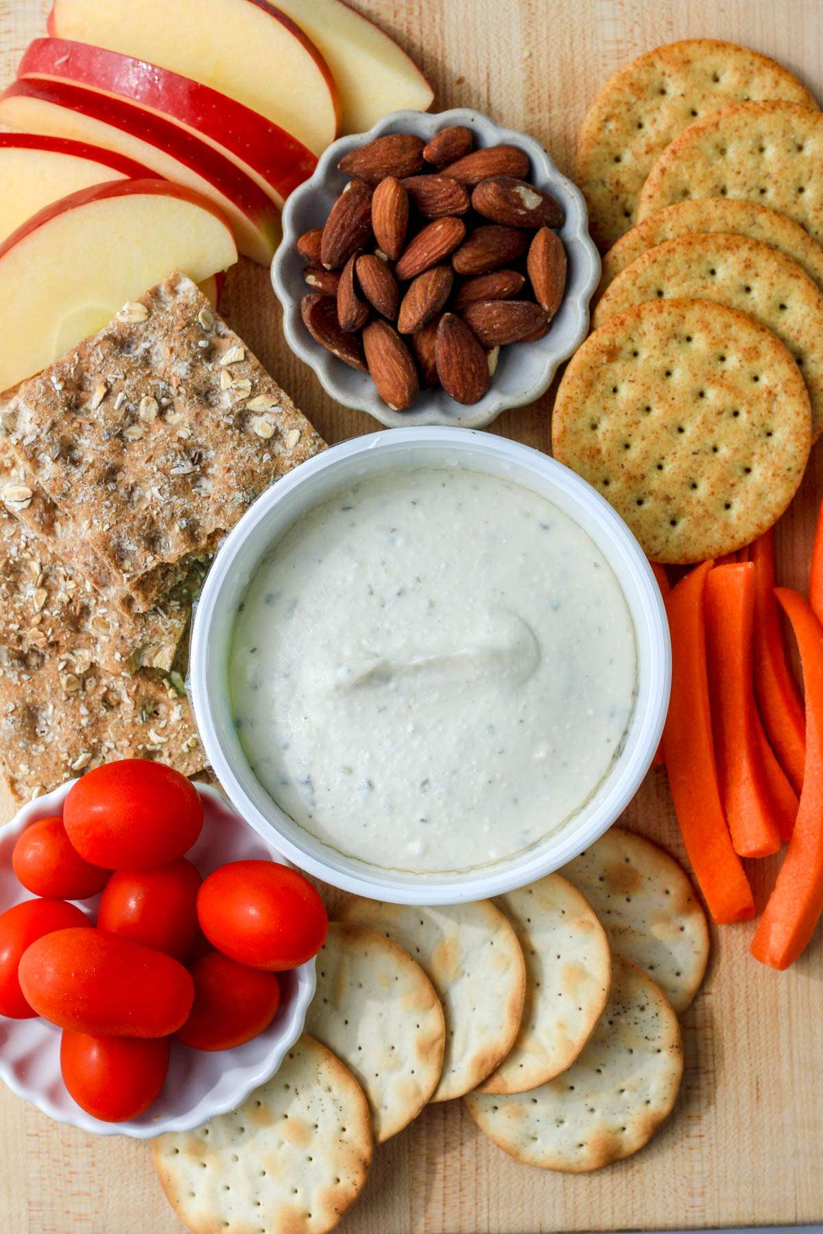 A wooden cutting board with various crackers, fruits, and vegetables surrounding dairy-free boursin cheese.