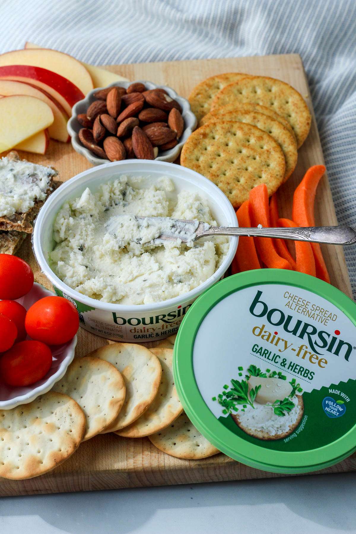 A wooden cutting board set up with crackers, veggies, fruit, and dairy-free boursin cheese with a silver spreader.